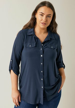 Camisa de botones azul marino con mangas enrolladas, dos bolsillos en el pecho y botones metálicos, fabricada en una tela suave y ligera.
