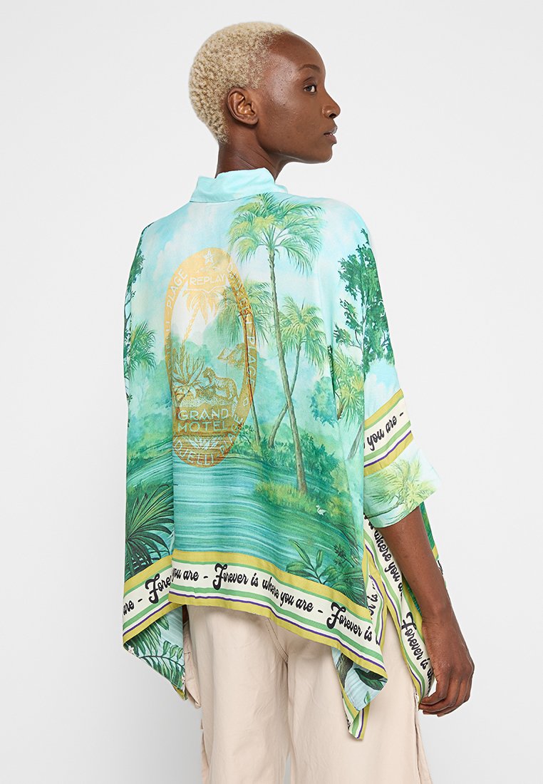 Osoba z krótkimi blond włosami, ubrana w szal w niebiesko-zielony tropikalny wzór, z palmami, wodą i napisem wzdłuż krawędzi.