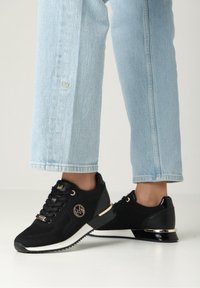 Schwarze Strick-Sneaker mit strukturiertem Obermaterial, weißer Sohle und goldenen Akzenten. Verfügt über ein Logo an der Seite und ein Detail mit schwarzem Lackleder an der Ferse.