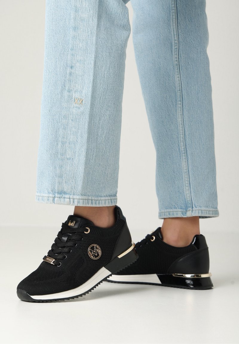 Schwarze Strick-Sneaker mit strukturiertem Obermaterial, weißer Sohle und goldenen Akzenten. Verfügt über ein Logo an der Seite und ein Detail mit schwarzem Lackleder an der Ferse.