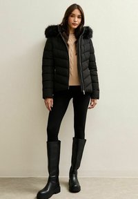 Kobieta ubrana w czarną kurtkę puchową z futrzanym kapturem, beżowy sweter, czarne legginsy i wysokie czarne botki, stojąca przy gładkiej ścianie.