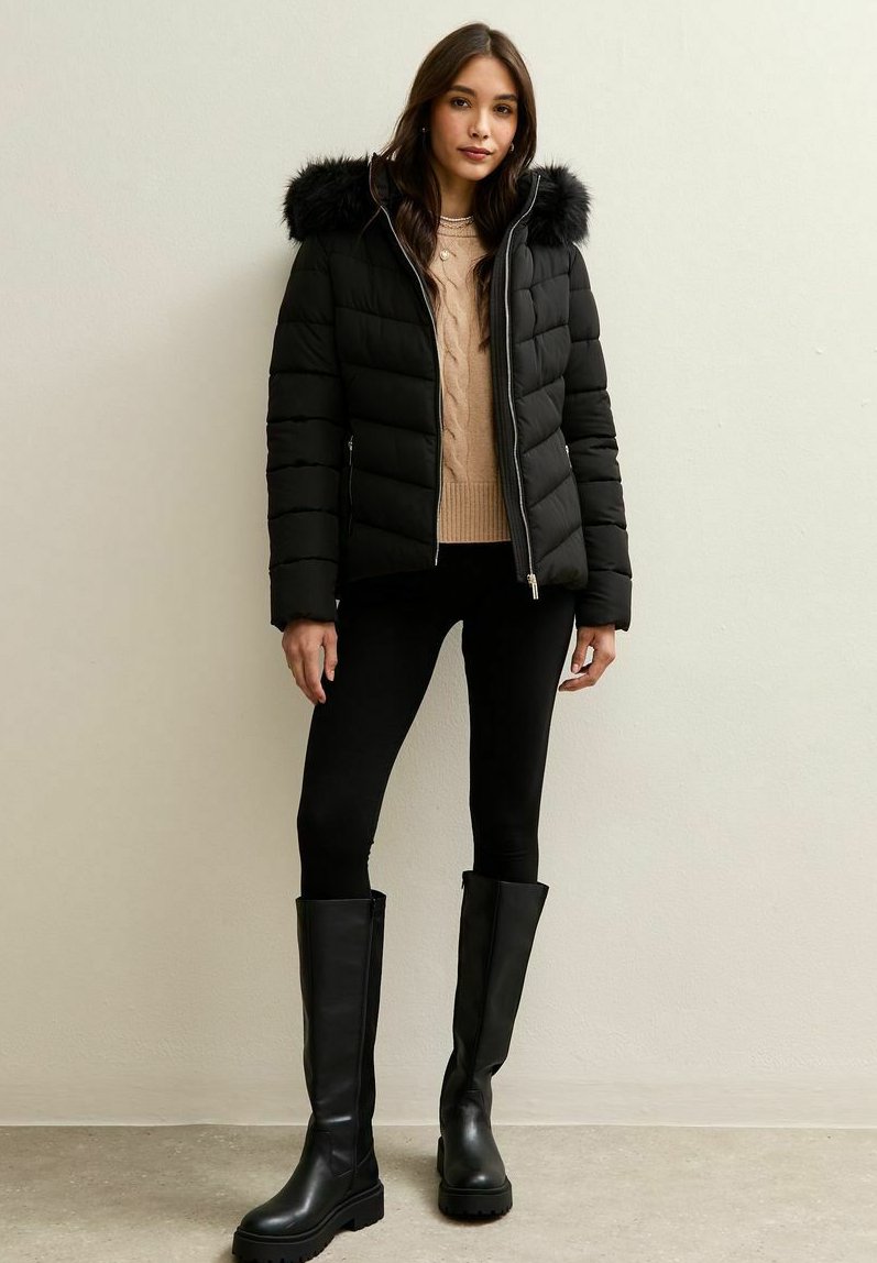 Kobieta ubrana w czarną kurtkę puchową z futrzanym kapturem, beżowy sweter, czarne legginsy i wysokie czarne botki, stojąca przy gładkiej ścianie.
