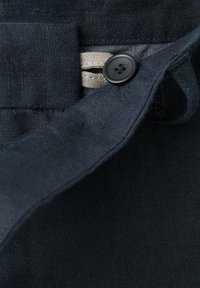 Gros plan d'un bouton en tissu foncé fixé à travers une boutonnière noire à quatre trous sur un vêtement avec des coutures visibles.