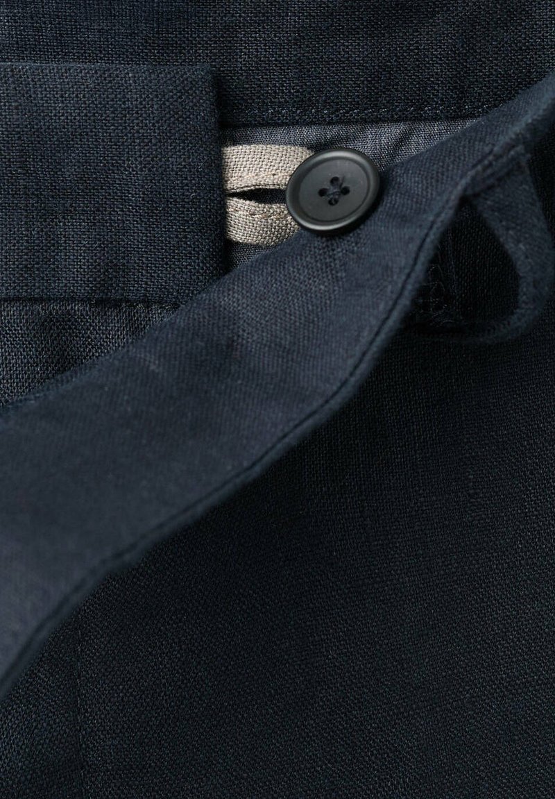 Gros plan d'un bouton en tissu foncé fixé à travers une boutonnière noire à quatre trous sur un vêtement avec des coutures visibles.