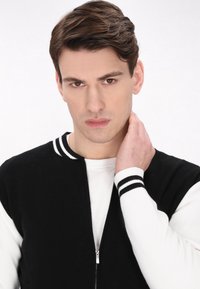 Juoda ir balta užtrauktuku užsegama striukė su dryžuotu megztu apykakle ir rankovėmis. Modelis liečia kaklą, vilkintis baltą marškinėlį po ja. Lygi tekstūra.