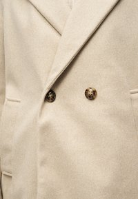 Manteau en laine beige avec larges revers, deux boutons visibles marron foncé et poches fendues de chaque côté.