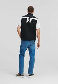 Svart ärmlös väst med vitt grafiskt mönster på baksidan, kombinerad med ljusblå jeans och svarta sneakers. Lätt tyg med en strukturerad passform.