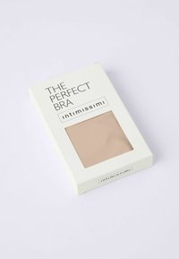 Naken bh förpackad i en vit låda märkt "DEN PERFEKTA BH:n", med ett transparent fönster för att visa produkten inuti.