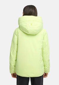 Vrouw in een lichtgroene gewatteerde jas met grote capuchon, gezien van achteren tegen een eenvoudige witte achtergrond.