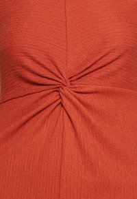 Haut couleur rouille en tissu texturé, avec un détail de nœud froncé au centre et de subtiles lignes verticales sur l'ensemble.