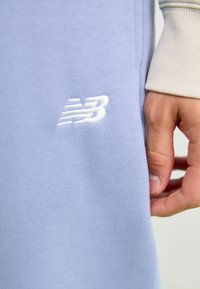 Hanorac de culoare mov deschis, realizat dintr-un amestec de bumbac, cu manșete ribbate și un logo brodat alb New Balance pe piept.