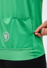 Χέρι που ανοίγει ένα μικρό μεταλλικό φερμουάρ στην πίσω τσέπη μιας υφασμάτινης πράσινης ποδηλατικής μπλούζας πάνω από μαύρο σορτς.