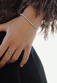 Srebrna narukvica s okruglim ukrasima, postavljena na ruci s tankim srebrnim prstenom s centralnim kamenom. Crna tkanina u pozadini.