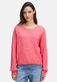 Frau in einem locker sitzenden pinken Pullover mit langen Ärmeln und hellblauen Jeans vor einem einfarbigen Hintergrund.