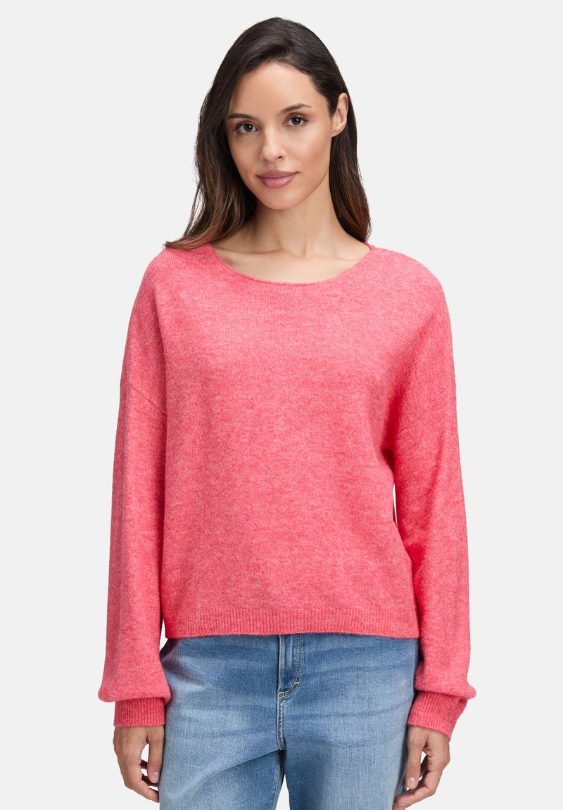 Frau in einem locker sitzenden pinken Pullover mit langen Ärmeln und hellblauen Jeans vor einem einfarbigen Hintergrund.