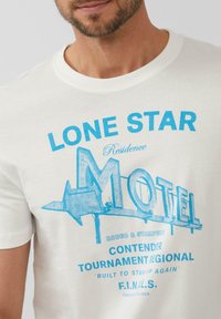 Crèmekleurig katoenen t-shirt met een blauw grafisch ontwerp van een motelbord met tekst, waaronder "LONE STAR" en "BUILT TO STAND UP AGAIN."