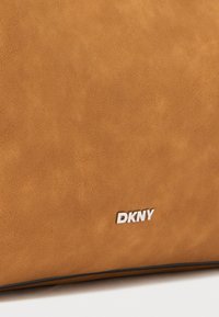 Tan leren tas met een gestructureerde afwerking, een zwarte rand en een zilveren DKNY-logo detail aan de zijkant.
