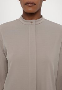 Lys taupe-bluse med ståkrage, som har en frontknaplukning, glat tekstur og minimalistisk design.