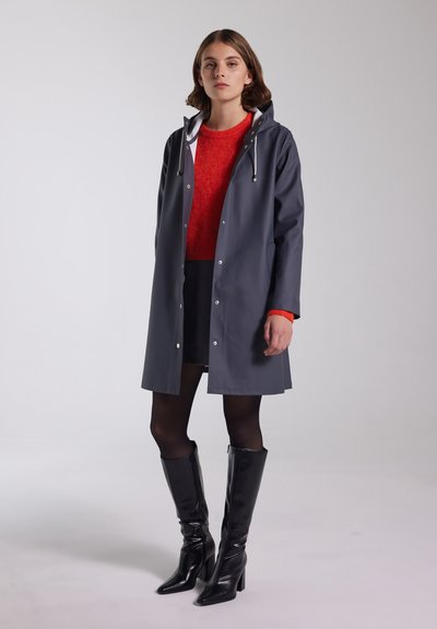 Grå vandtæt regnfrakke med hætte, trykknapper, hvid foring, båret over en rød sweater og sort nederdel, parret med sorte knæhøje støvler.