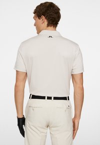 Ljusbeige pikétröja med krage, korta ärmar och svart logodetalj på baksidan. Bärs tillsammans med krämfärgade byxor och ett svart bälte.