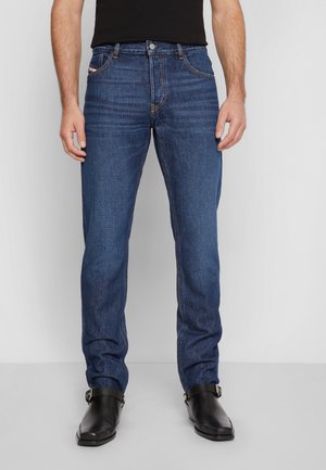 Mann trägt Slim-Fit blaue Jeans und schwarze Lederstiefeletten, steht vor einem schlichten hellgrauen Hintergrund.