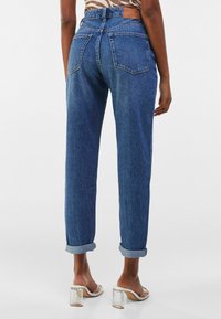 Vaqueros de mezclilla azul oscuro con un corte relajado, puños enrollados y dos bolsillos traseros. Presenta un parche de cuero marrón en la cinturilla.