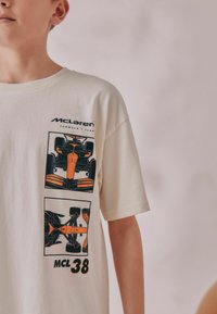 Cremefarvet t-shirt med McLaren Formel 1 grafik i sort og orange, der fremviser bildesigns og teksten "MCL 38". Korte ærmer.