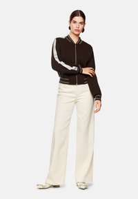 Braune Bomberjacke mit Reißverschluss und kontrastierendem grauen Ärmelleisten; getragen über weißen, weiten Hosen. Sie verfügt über einen gerippten Kragen und feine Nähte.