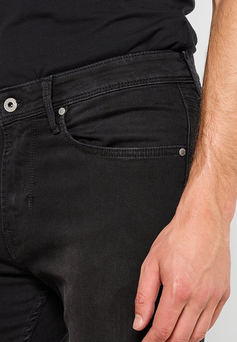 Jeans de mezclilla negra con corte ajustado. Las características incluyen cinco bolsillos, costuras visibles y un botón de metal en la cintura.