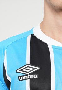 Camiseta deportiva a rayas azul claro y negra con acentos blancos y un logo negro de "Umbro" en el pecho. Textura de tejido suave, diseño moderno.