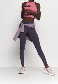 Różowy krótki top sportowy z logo Nike, w połączeniu z fioletowymi legginsami z wysokim stanem. Na talii związany jasnofioletowy sweter i różowe buty.