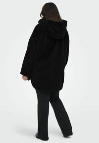 Liela izmēra melna flīsa jaka ar kapuci, ar mīkstu tekstūru. Modelis redzams no aizmugures, ģērbts melnās biksēs un apavos.