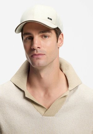 Homme portant un pull en maille texturée beige clair avec col ouvert et une casquette crème avec le logo BOSS sur fond blanc.