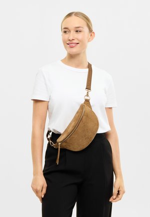 Riñonera de ante marrón con forma curva, cremallera dorada y correa desmontable. Combinada con una camiseta blanca y pantalones negros.