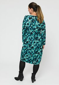 Šaty s dlhými rukávmi v odtieni teal a čiernej s kruhovým bodkovaným vzorom, priliehavé v oblasti pása a jemne sa rozširujúce na leme. Nosené s čiernymi členkovými topánkami.