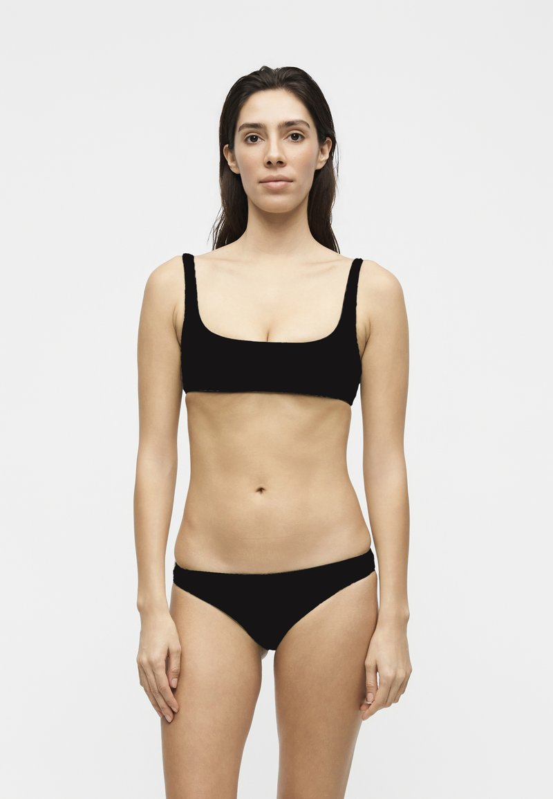 Vrouw met lang donker haar die een zwarte bikini draagt, rechtopstaand tegen een eenvoudige lichtgrijze achtergrond.
