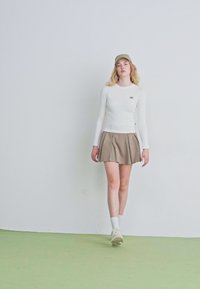 Bela dolga majica z dolgimi rokavi, bež nabran krožnik, in svetlo zelena kapa. Model nosi bele nogavice in bež športne copate, stoji na svetlo zeleni tleh.