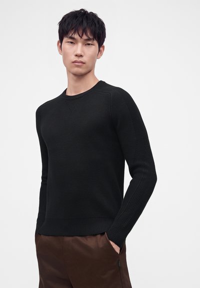 Jeune homme aux cheveux courts noirs portant un pull en maille noir et un pantalon marron, debout avec une main dans la poche sur fond blanc.