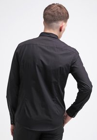 Musta pitkähihaisi paita, jossa on mukautuva malli, napitettavat hihansuut, sileä pinta ja hienovarainen kiilto, nähtynä takaa.