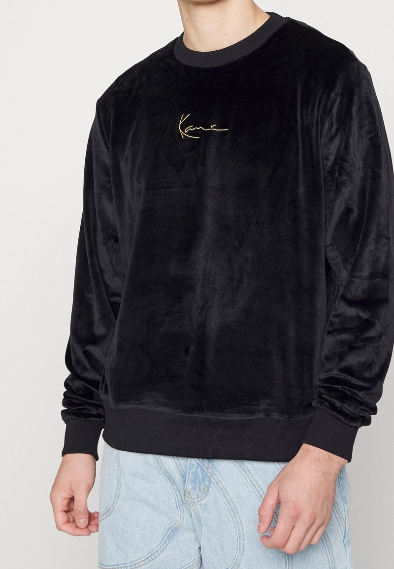 Sweat-shirt noir en velours à manches longues avec un petit logo doré brodé sur la poitrine, porté avec un jean bleu clair.
