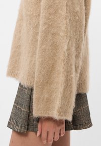 Mão repousada ao lado de uma saia curta de xadrez castanho, parcialmente coberta pela manga de um suéter bege felpudo, contra um fundo claro e simples.