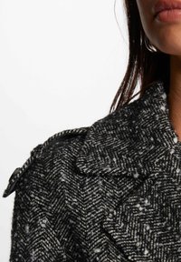 Zwart-wit houndstooth wollen jas met grote revers, textuurstof en een knoopdetail op de schouder.