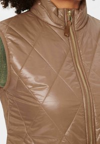 Brązowa, pikowana kamizelka z wysokim kołnierzem, metalowym zamkiem i błyszczącą teksturą. Posiada zielone dzianinowe akcenty w okolicy ramion.