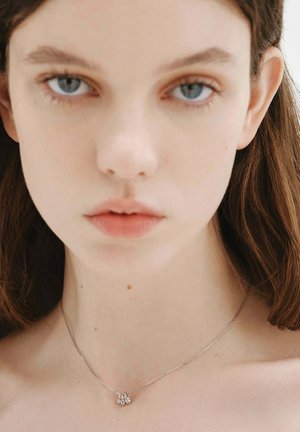 Joven con cabello castaño liso que lleva un delicado collar de plata con tres pequeños colgantes de piedra preciosa transparente.