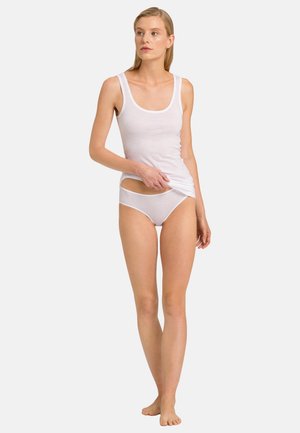 Woman wearing white tank top and matching white underwear, standing barefoot against a plain light background.