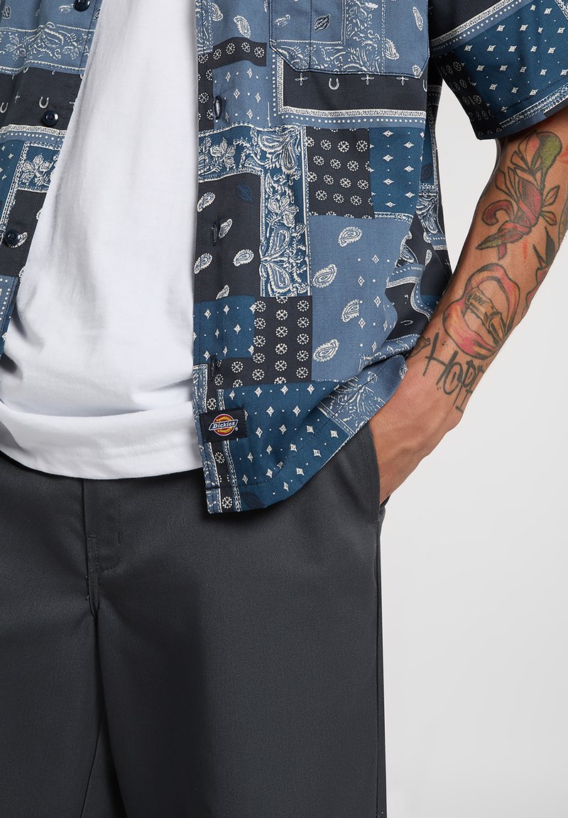 Camicia con bottoni caratterizzata da un motivo bandana blu con accenti scuri a contrasto, abbinata a pantaloni grigi. Etichetta visibile all'orlo.