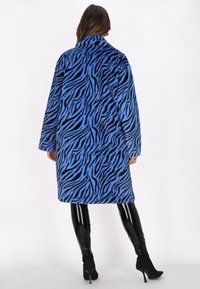 Cappotto blu fuzzy con strisce di tigre nere, colletto alto, maniche lunghe e vestibilità rilassata, abbinato a leggings neri lucidi e stivaletti.
