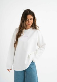Witte longsleeve sweatshirt met paarse tekst "MIRONS" gecentreerd op de borst, met een relaxte pasvorm en ronde halslijn. Spijkerbroek zichtbaar eronder.