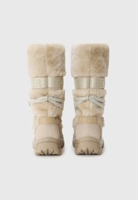 Bézs szőrrel bélelt csizmák vastag talppal, "BO" feliratos pántokkal és dekoratív masnival a hátulján.