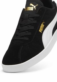 Fekete velúr sneaker fehér oldalsó csíkkal és arany Puma logóval. Kerek fűzőkkel és texturált fehér gumitalppal rendelkezik.